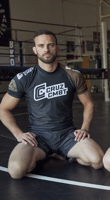 Man sitting on a boxing ring wearing a Cruz CMBT shirt in a gym setting