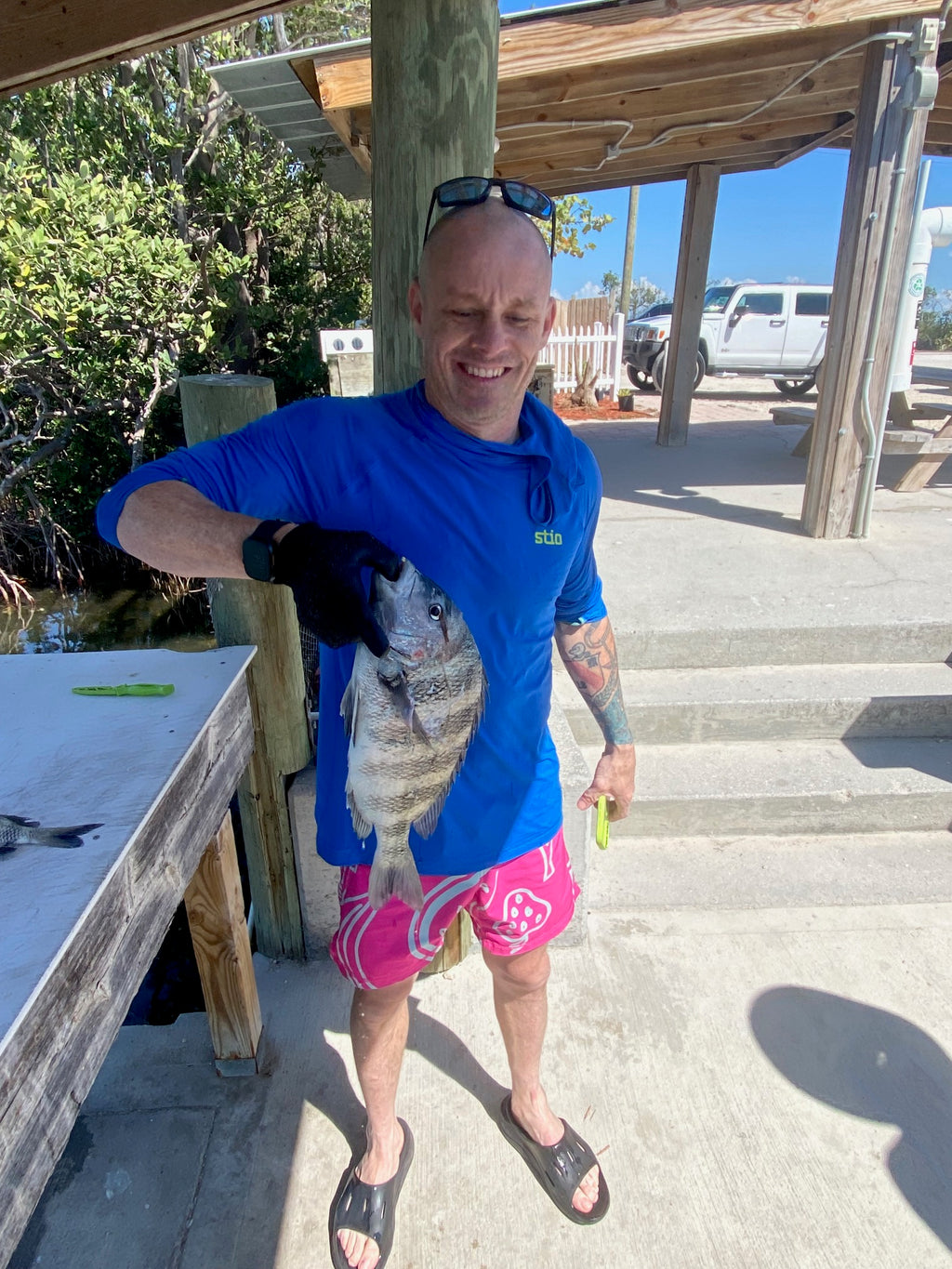 Man holding a large fish near a wooden structure with a concrete wall in the background.