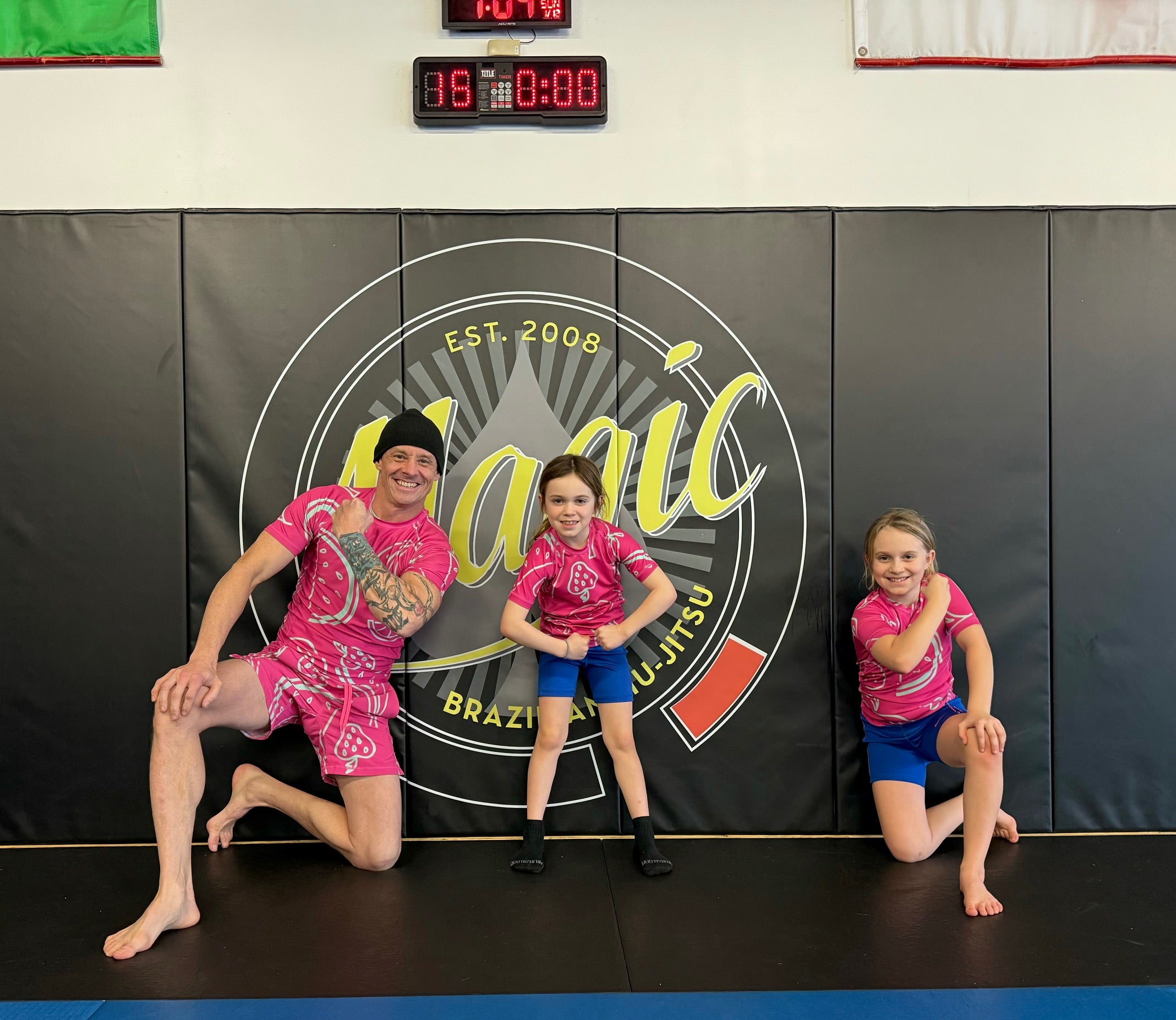 Three people in pink shirts posing in front of a Brazilian Jiu-Jitsu logo.