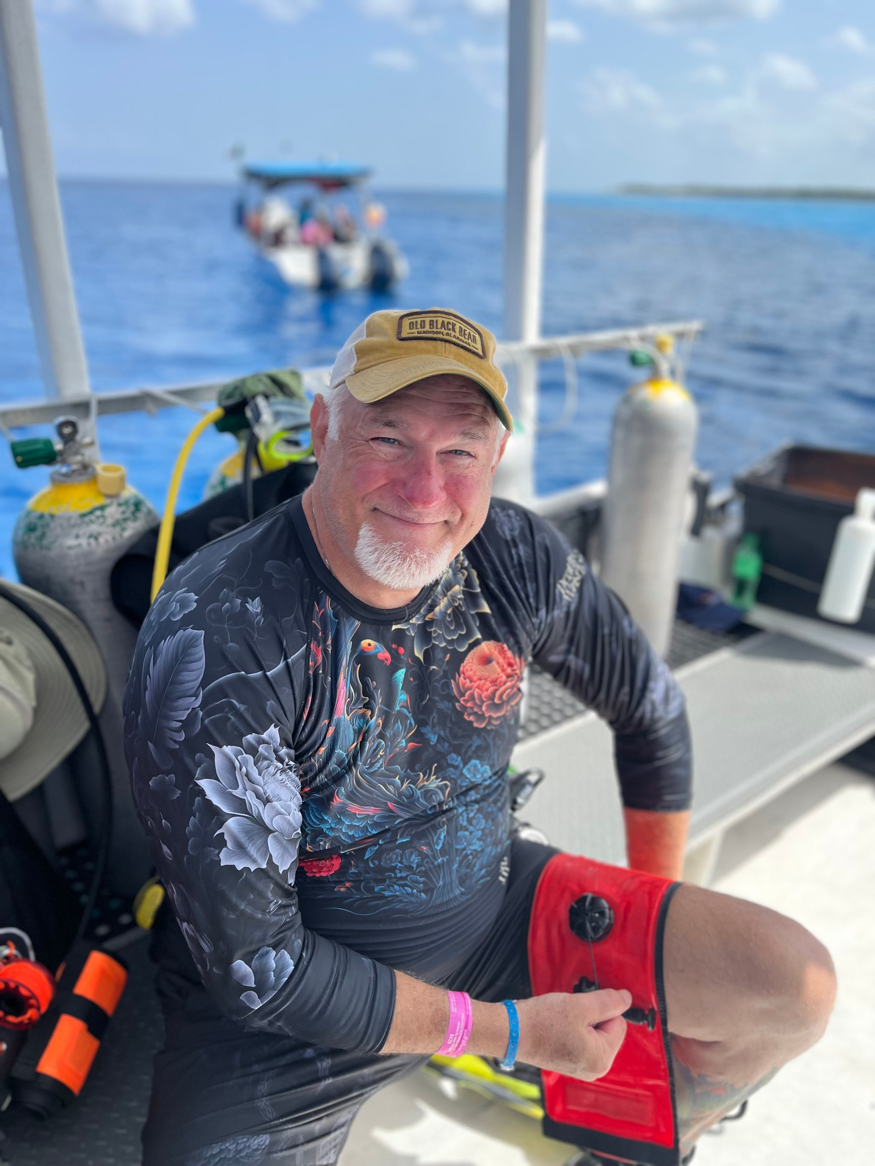 Man on a boat with diving equipment, wearing a cap and wetsuit.