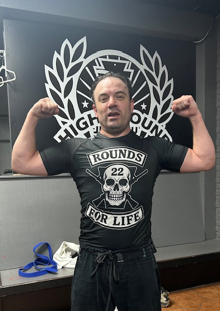 Man flexing muscles in a gym wearing a 'Rounds for Life' t-shirt.